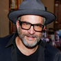 Smiling man with a beard and glasses wearing a dark fedora indoors.