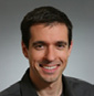 Head-and-shoulders portrait of a smiling man with short dark hair wearing a dark blazer.