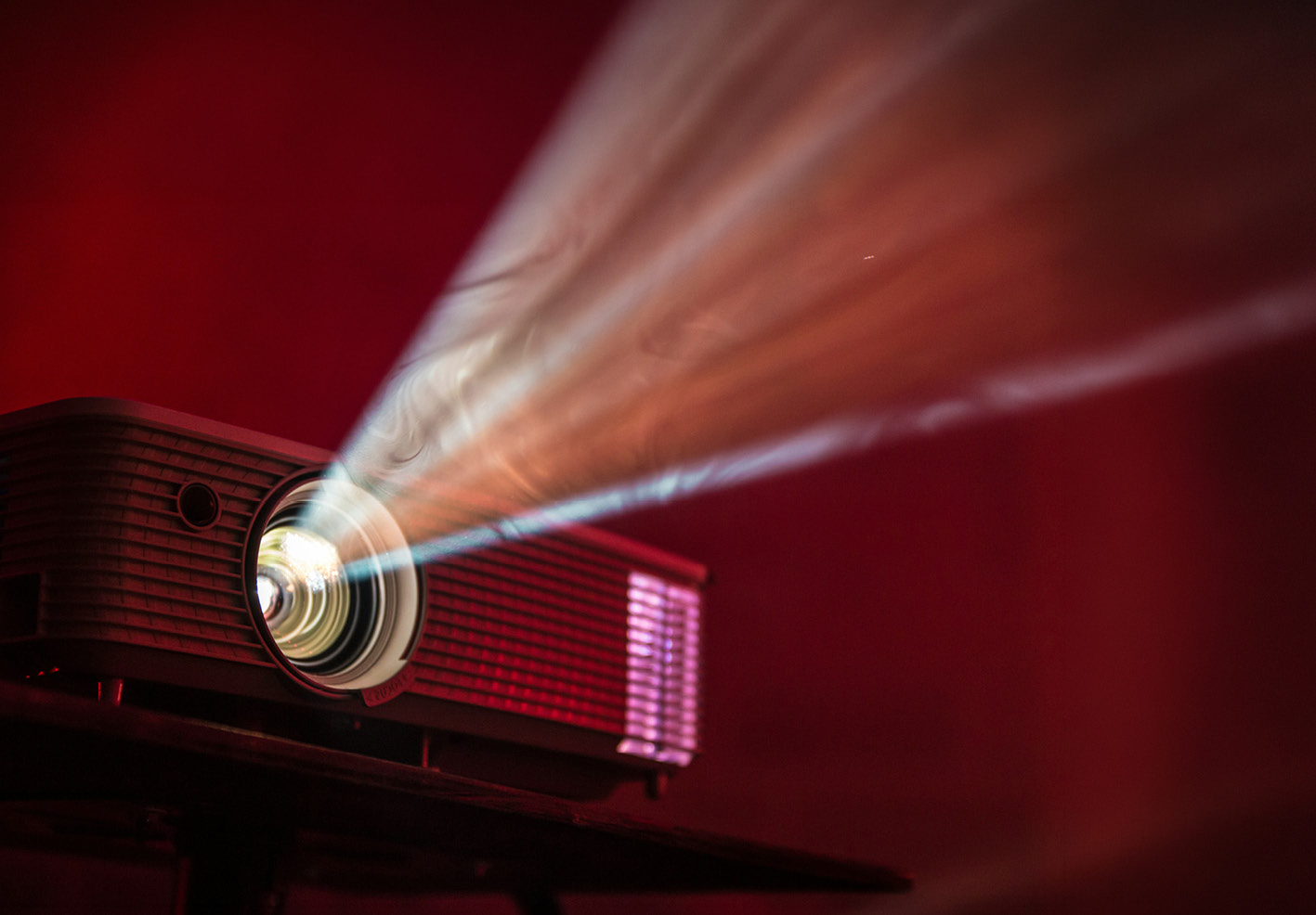 A projector casting a bright beam of light across a dark red room, with the lens glowing.