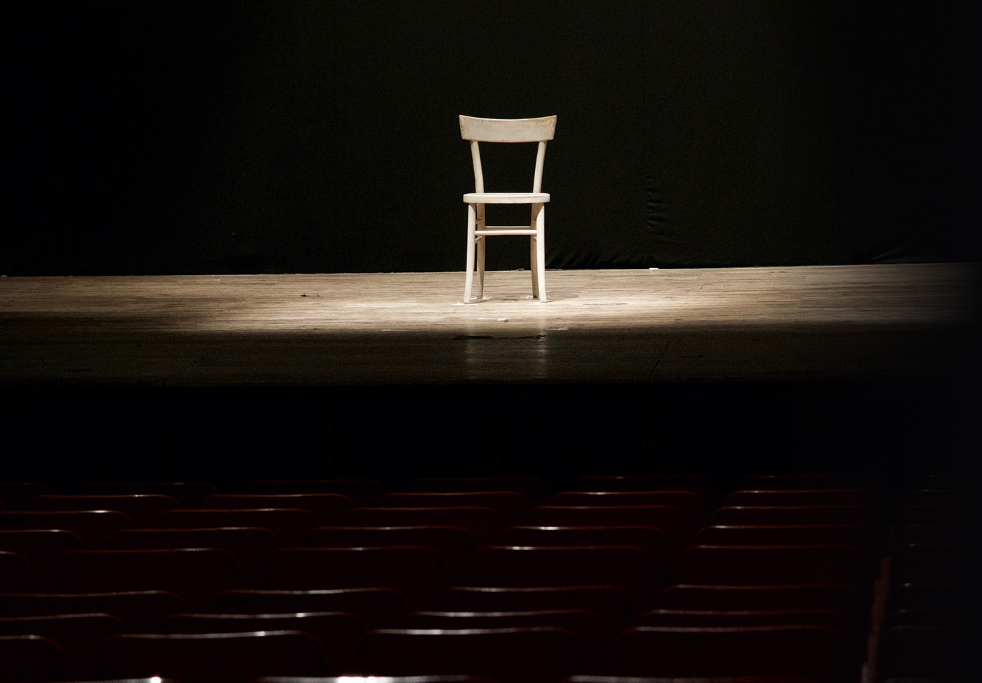 A lone white wooden chair centered on a spotlighted stage in a dark theater.