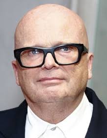 Bald man wearing black-framed glasses and a dark suit, looking at the camera.