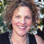 Portrait of a smiling woman with curly brown hair, outdoors among trees in daylight.