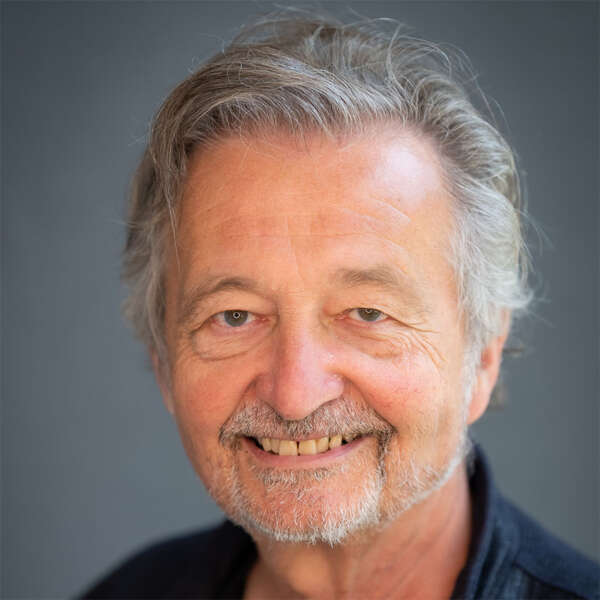 Close-up portrait of a smiling older man with gray hair and light stubble against a neutral gray background.